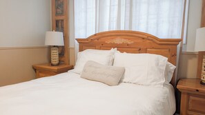 Una cama bien tendida con sábanas blancas y un cojín con texto, un cabecero de madera, una mesita de noche con una lámpara, y una ventana con cortinas traslúcidas.