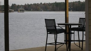 Une terrasse en bois avec deux chaises et une table, donnant sur un lac, avec des bâtiments et des arbres en arrière-plan.