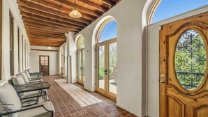 A corridor with a tiled floor, wooden ceiling beams, arched windows, and a wooden door with a decorative glass panel.