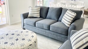 A living room with a grey sofa, striped pillows, a round ottoman with nautical prints, and a dining area with white chairs and a ship's wheel decoration. - Beachfront Dream Cottage Sleeps 11 Beautiful PETS OK (Ocean City)