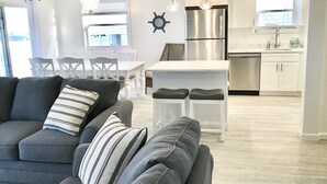 A modern living room with a grey sofa and striped pillows, a dining area with white chairs, and a kitchen with stainless steel appliances and white cabinetry. - Beachfront Dream Cottage Sleeps 11 Beautiful PETS OK (Ocean City)