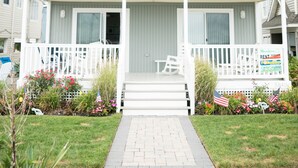 A two-story building with a white exterior, a front porch, and a small garden with American flags. - Beachfront Dream Cottage Sleeps 11 Beautiful PETS OK (Ocean City)