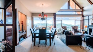 A spacious living room with a large dining table, comfortable seating, a chandelier, and large windows offering a view of the outdoors. - Altitude - Tremblant Luxurious Ski-in Ski-out Condo - Breathtaking Views (Mont-Tremblant)