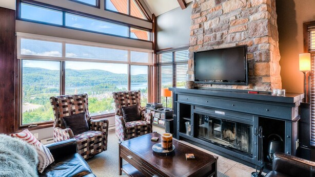 A cozy living room with a stone fireplace, large windows, and comfortable seating. - Altitude - Tremblant Luxurious Ski-in Ski-out Condo - Breathtaking Views (Mont-Tremblant)