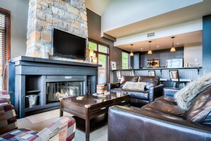 A modern living room with a stone fireplace, a flat-screen TV, leather sofas, and a kitchen area with pendant lighting. - Altitude - Tremblant Luxurious Ski-in Ski-out Condo - Breathtaking Views (Mont-Tremblant)