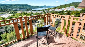 A wooden deck with a glass table and chairs overlooking a scenic landscape with hills and a lake. - Altitude - Tremblant Luxurious Ski-in Ski-out Condo - Breathtaking Views (Mont-Tremblant)