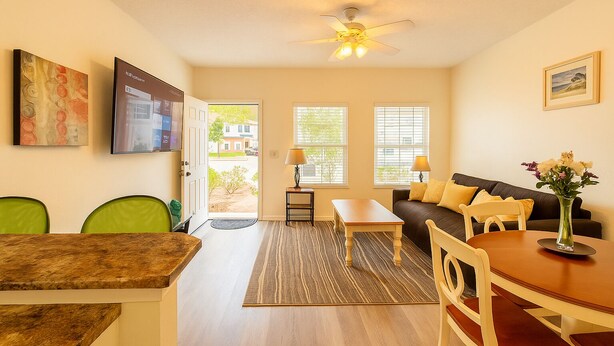 A living room with a sofa, a coffee table, and a dining table with chairs. There is a television mounted on the wall and a ceiling fan.