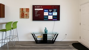 A modern living room with a flat-screen TV mounted on the wall, a glass-top coffee table with a sculpture, green chairs, and a patterned rug.