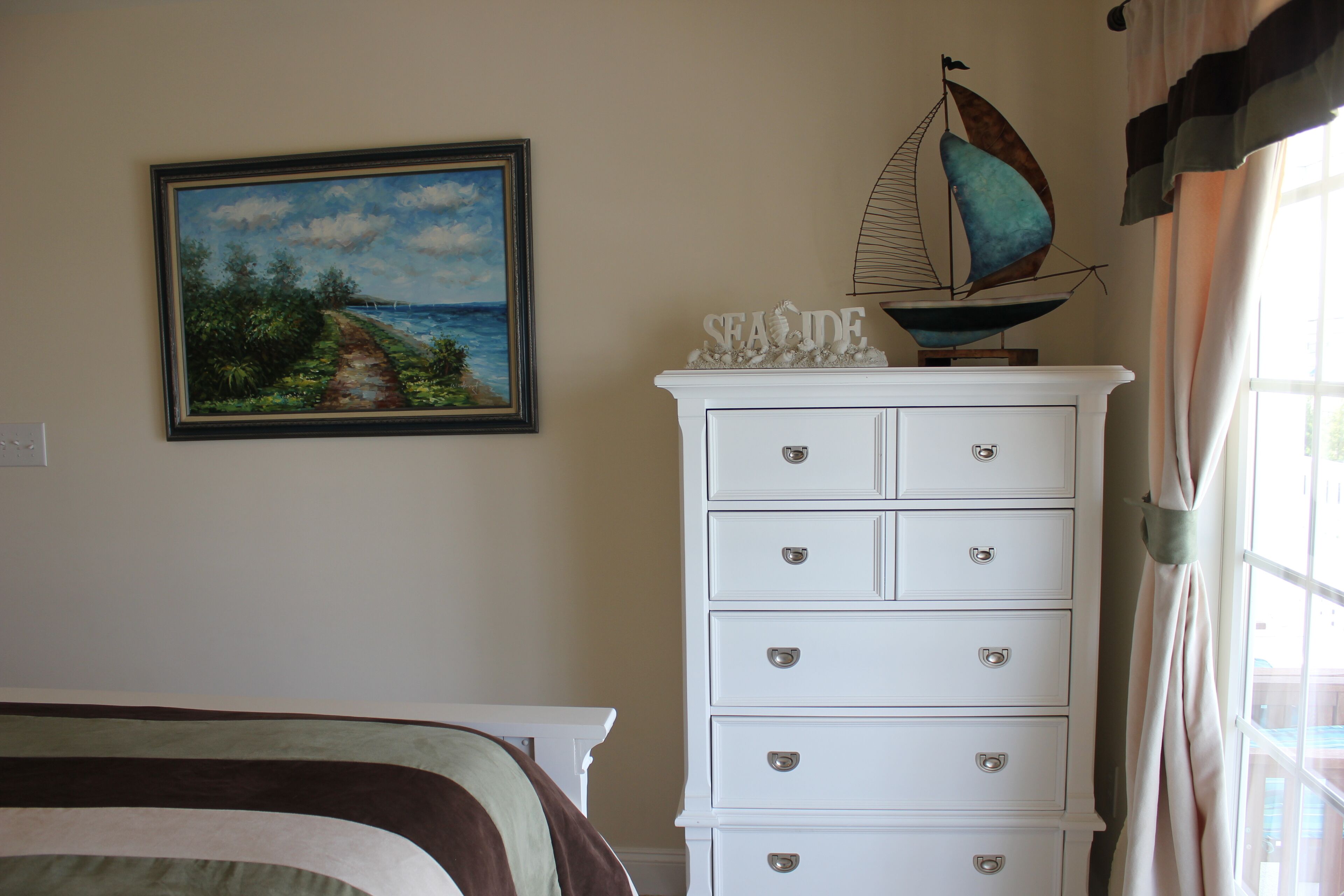Une chambre à coucher comprenant un lit, une commode avec des tiroirs, un tableau représentant un paysage côtier et une décoration sur le thème d’un voilier.