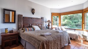 Quarto com cama grande, mesa de cabeceira, cabeceira de madeira, espelho e janela com vista para vegetação.