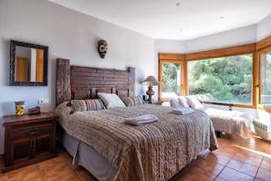 A bedroom with a large bed, a nightstand, a wooden headboard, a mirror, and a window with a view of greenery.