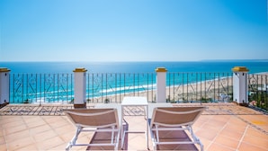 Terraço com duas cadeiras brancas e uma mesa, com vista para uma praia de águas azuis cristalinas e areal dourado.