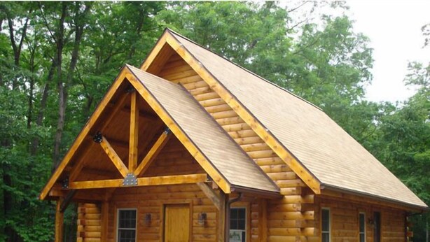 Eine Blockhütte mit markantem Satteldach, einer Veranda mit Geländern und einem kleinen Vorgarten mit Sträuchern.