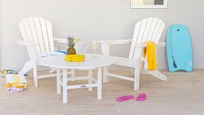 Une table blanche avec deux chaises Adirondack blanches, un ananas, un verre de vin blanc et une serviette jaune posée sur la table.