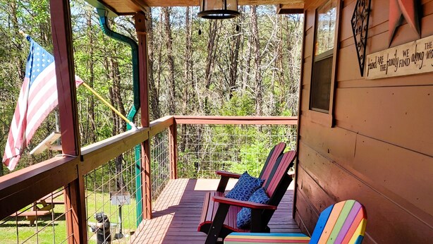 A covered deck with wooden flooring, a ceiling fan, and a view of a forest. There are two Adirondack chairs with colorful cushions. - Private Hideaway near Pigeon Forge; Convenient to Attractions; Five-Star Reviews (Sevierville)
