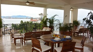A spacious balcony with a view of the city and the sea, furnished with wooden chairs and a round table with a fruit bowl and cups. - Beachfront Panoramic Oceanview Modern Luxury (Old Town Romantic Zone) (Puerto Vallarta)