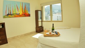 A hotel room with a bed, a painting of sailboats, a window with a city view, and a tray with a bottle and glasses. - Beachfront Panoramic Oceanview Modern Luxury (Old Town Romantic Zone) (Puerto Vallarta)