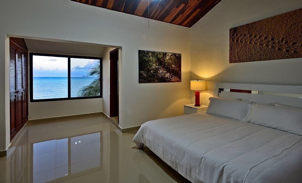 A bedroom with a large window, a bed, a nightstand, and a wall-mounted artwork.