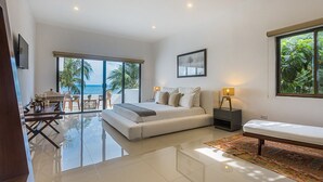 Une chambre moderne avec un grand lit, une table de chevet avec une lampe, un banc et une vue sur l’océan grâce à de grandes fenêtres.