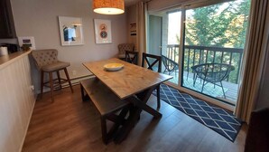 A dining area with a wooden table, chairs, and a bowl. There is a sliding glass door leading to a balcony with outdoor furniture.