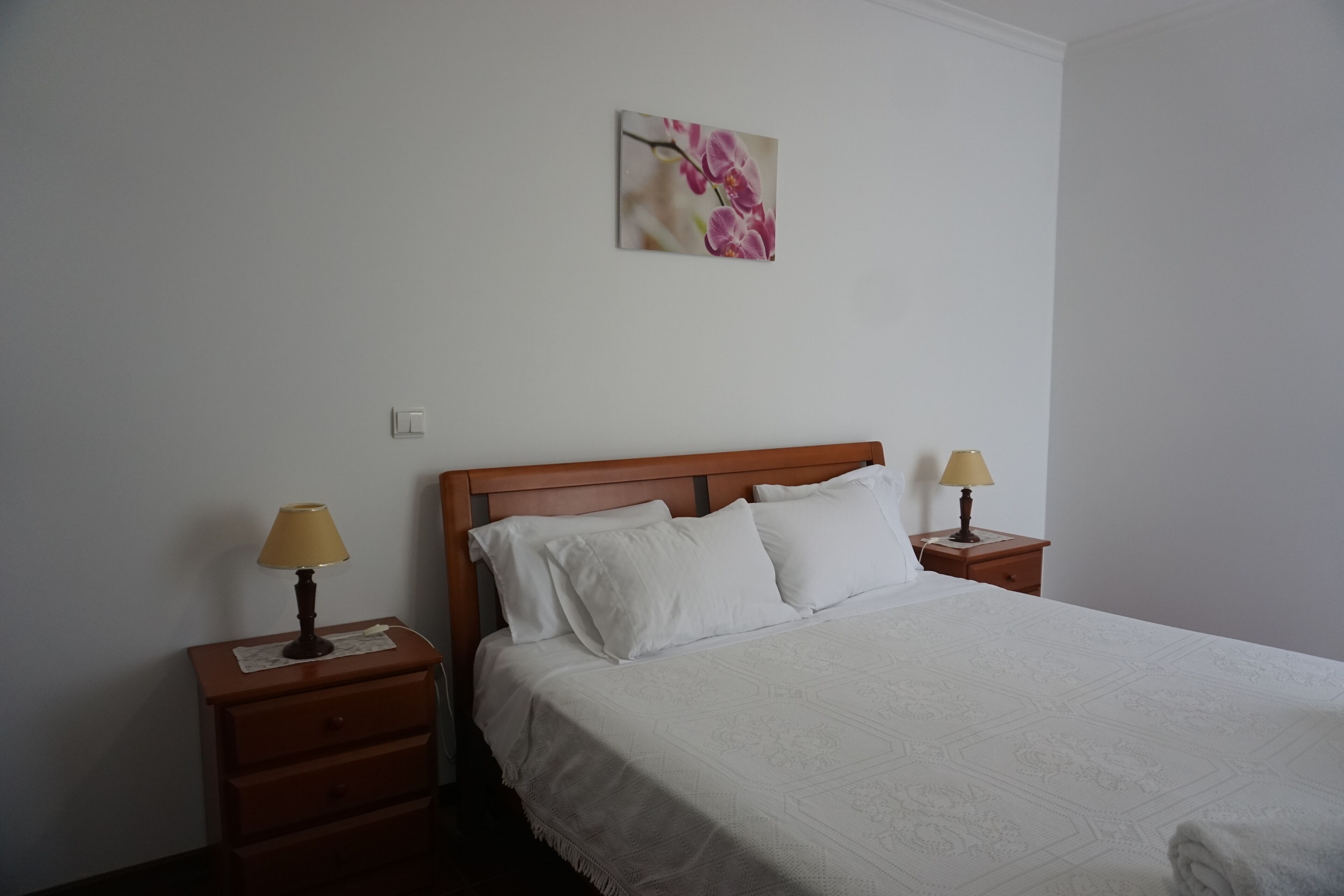 Una cama bien hecha con ropa blanca, dos mesitas de noche con lámparas y un cuadro de flores colgado en la pared.