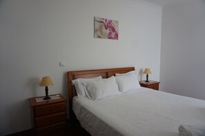 Ein ordentlich gemachtes Bett mit weißer Bettwäsche, zwei Nachttische mit Lampen und ein an der Wand hängendes Bild von Blumen.