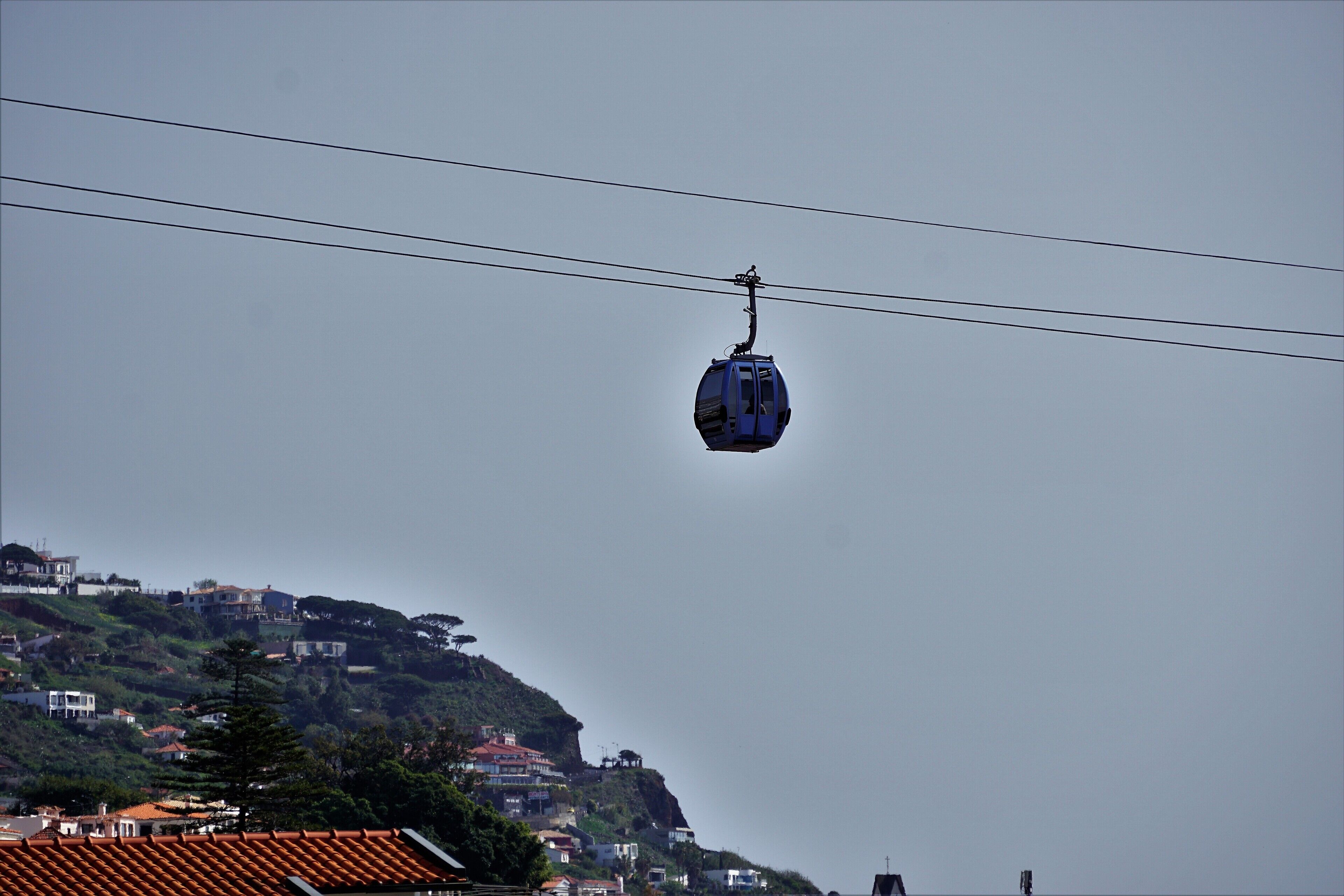 A cable car suspended between two power lines, with a hillside of buildings in the background.