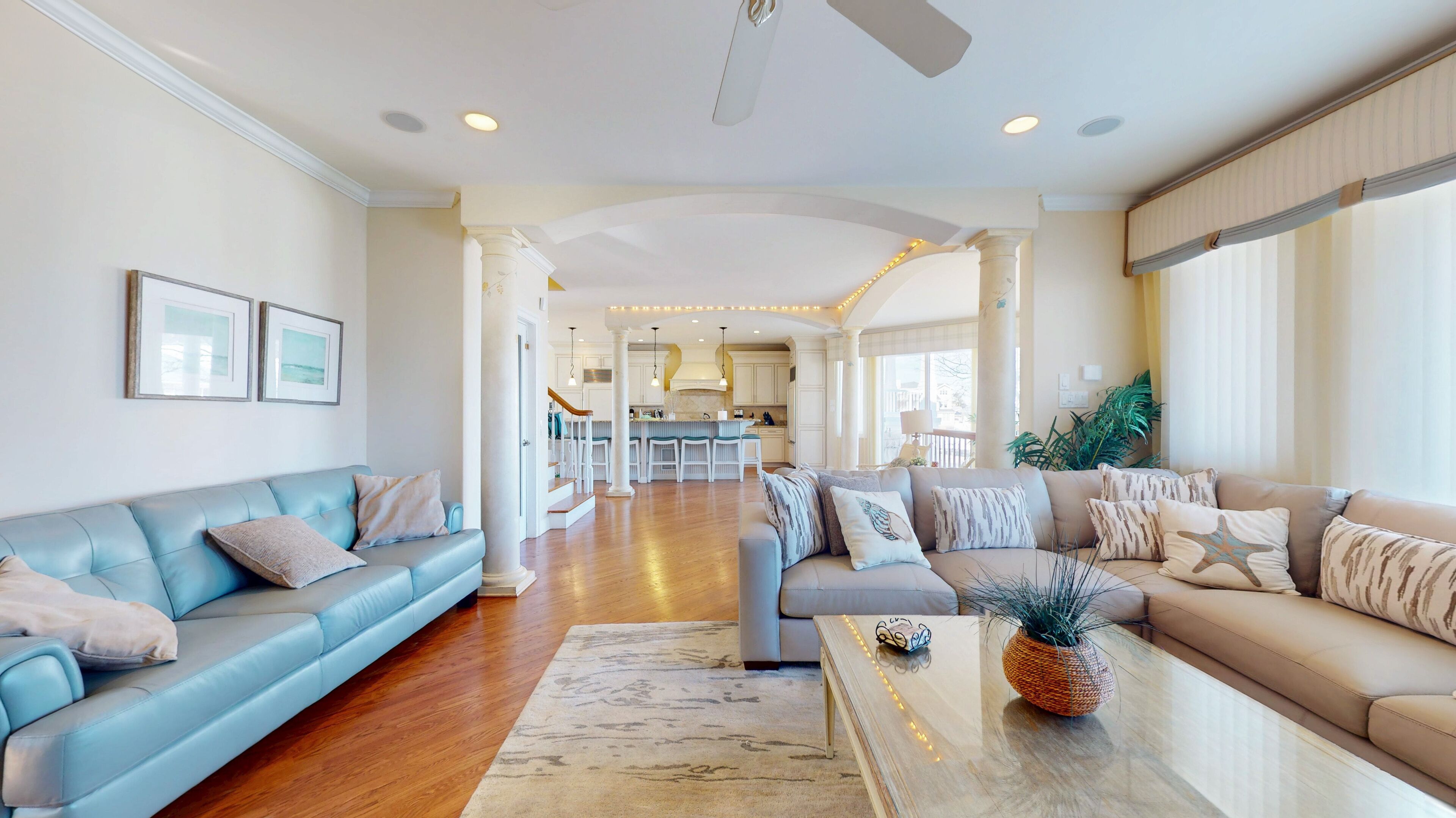 A spacious living room with a light blue sofa, a beige sofa, and a coffee table with a plant. A kitchen is visible in the background.