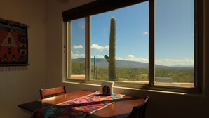 A dining area with a wooden table and chairs, a colorful table runner, and a window with a view of a cactus and mountains. - Amazing City And Mountain Views From The Foothills Of The Catalinas (Tucson)