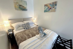 A neatly made bed with a patterned bedspread, a decorative pillow, and a towel on the side. There are wall art pieces above the bed and a nightstand with a lamp and a small box. - Heaven Can't Wait:  ON the BEACH!! Heated pool + hot tub, private dock. 7BR/5BA (Stuart)