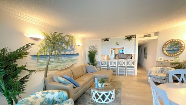 A living room with a beach mural, a sofa, a floral armchair, a coffee table, and a dining area with a white table and chairs.