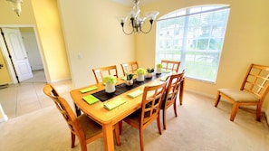 Comedor con una mesa de madera dispuesta para seis personas, rodeada de sillas, con una araña colgante y una ventana con persianas a la derecha.
