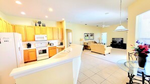 A modern kitchen with wooden cabinets, a white refrigerator, and a built-in oven. There is a dining area with a glass table and a black chair. A living room with a sofa and a TV is visible in the background. - Immaculate Spacious Villa.No rear neighbors,Games Room,office,Large pool,hot spa (Davenport)