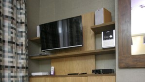 Een moderne woonkamer met een flatscreen televisie bevestigd aan een houten kast, een klein tafeltje met een metalen vaas, en een kamerbreed gordijn met een patroon.