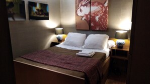 Een net opgemaakt bed met witte kussens en een opgevouwen handdoek, twee bedlampen, en een muur met een groot schilderij.