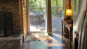 A spacious room with a stone fireplace, wooden ceiling beams, and a view of a deck with outdoor furniture. - Cozy Cabin Secluded in the Woods.  Wildlife Abundant.  Not a neighbor in sight! (Harshaw)