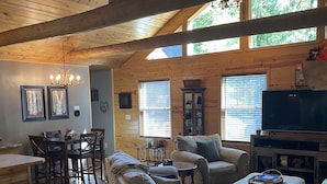 A spacious living room with a wooden ceiling, a dining area with a wooden table and chairs, and a cozy seating area with a sofa and ottoman. - Cozy Cabin Secluded in the Woods.  Wildlife Abundant.  Not a neighbor in sight! (Harshaw)