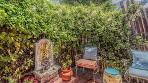 A garden with a stone fountain, a wooden chair with a cushion, and a potted plant.