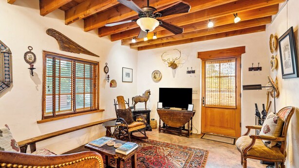 A rustic living room with wooden beams, a ceiling fan, a large window with blinds, a fireplace, and a television.