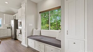Une cuisine moderne avec des meubles blancs, un banc intégré doté de coussins et une fenêtre donnant sur un espace verdoyant.