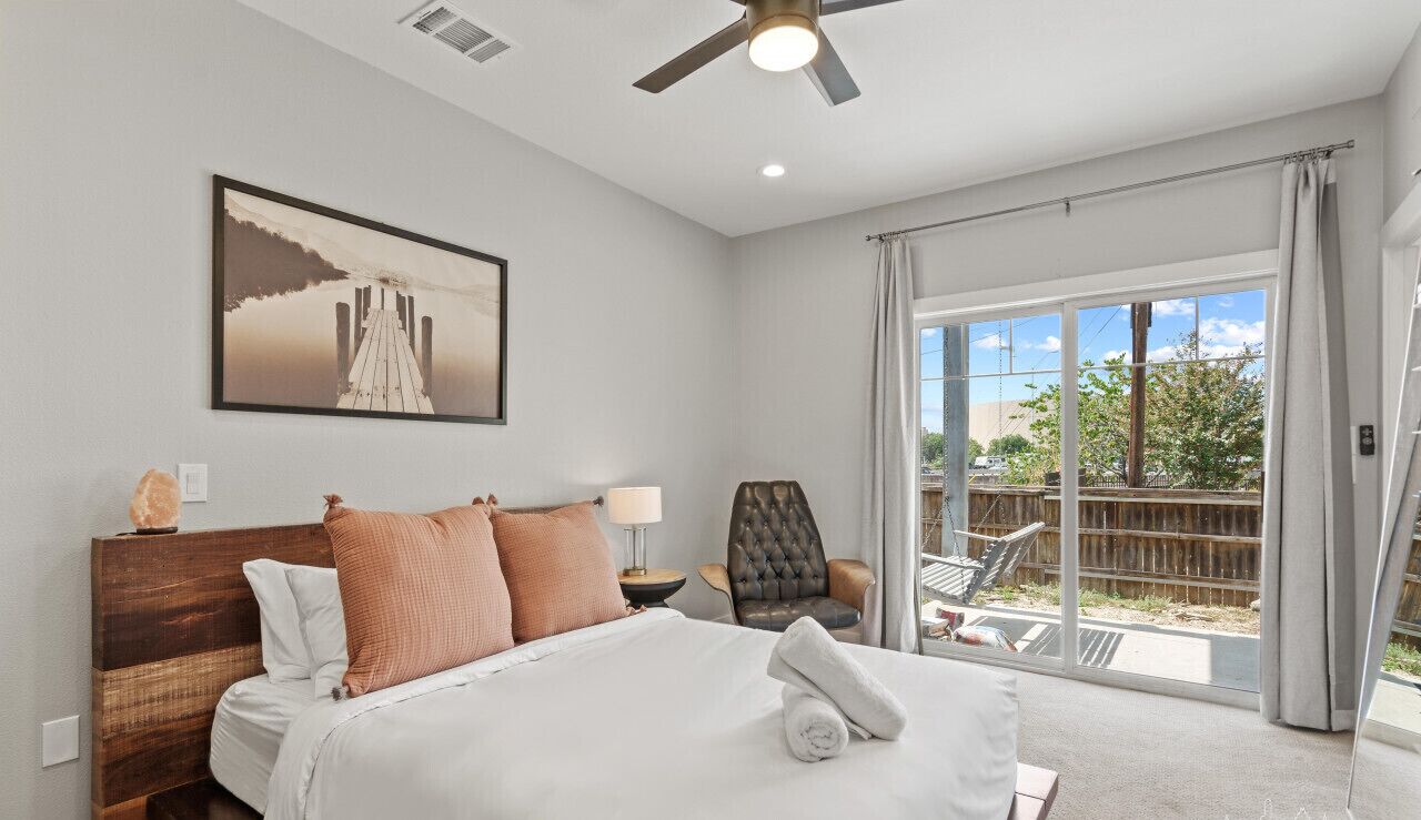A bedroom with a large bed, a ceiling fan, a nightstand, a chair, a window with curtains, and a painting on the wall.