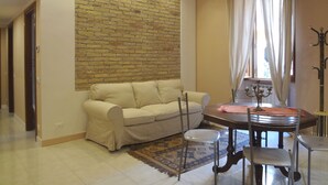 A living room with a beige sofa, a dining table with chairs, a patterned rug, and a brick accent wall. - Rome Historic Core Apartment - Between Colosseum & Termini – A/C, Wi-Fi, Netflix (Roma)