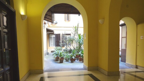 A corridor with yellow walls and a tiled floor, leading to a courtyard with potted plants and a view of buildings. - Rome Historic Core Apartment - Between Colosseum & Termini – A/C, Wi-Fi, Netflix (Roma)