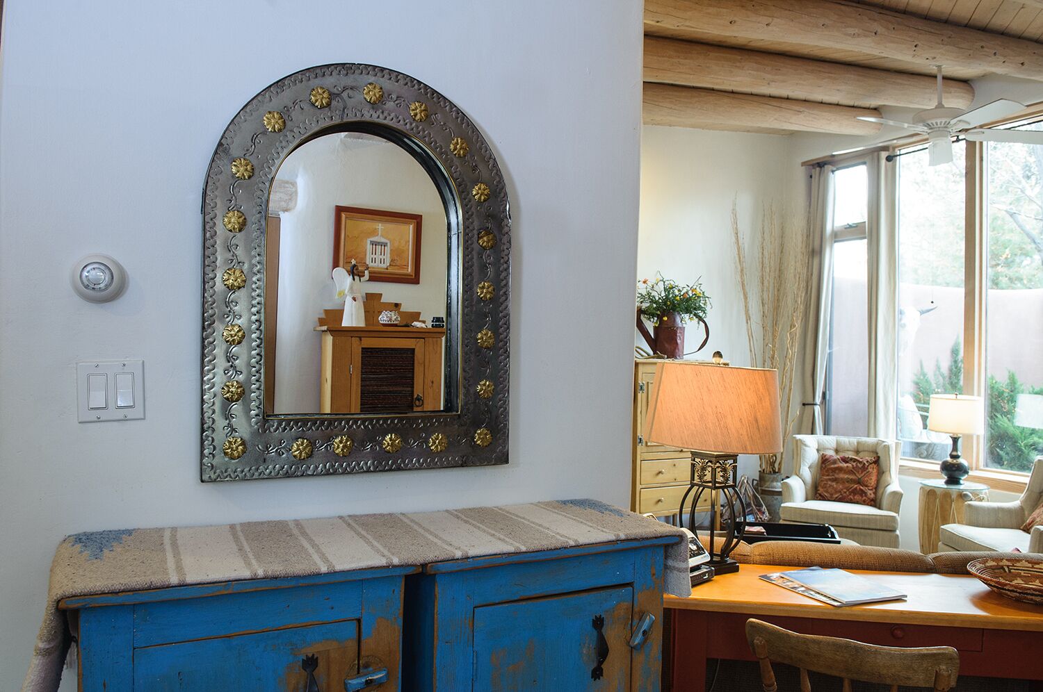 A room with a large mirror, a blue chest of drawers, a wooden table with a lamp, and a window with curtains.