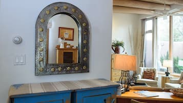 A room with a large mirror, a blue chest of drawers, a wooden table with a lamp, and a window with curtains.