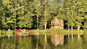 A lakeside cabin with a deck, surrounded by dense trees and reflecting on the water. - Lakefront 🛶 • Spa ♨️ • Firepit 🔥 • Boating 🚣‍♂️ • Game Rm 🎮 • Fishing 🎣 • Sunsets 🌅 (Poconos Lake)