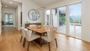 Un comedor amplio con una mesa de madera y sillas blancas, un espejo grande y vistas al exterior a través de puertas de cristal correderas.