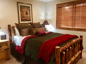 Quarto com estrutura de cama de madeira, colcha vermelha e verde, e cabeceira de madeira. Há uma mesa de cabeceira com um abajur e um quadro na parede.