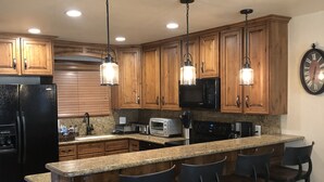 A modern kitchen with wooden cabinets, a black refrigerator, a microwave, a toaster, and a bar area with stools. - TRUE SKI IN/SKI OUT, SLOPESIDE 3 BR CONDO - PATIO, HOT TUB, POOL, FREE PARKING (Park City)