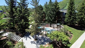 A pool area surrounded by tall trees, with a building in the background and a clear sky. - TRUE SKI IN/SKI OUT, SLOPESIDE 3 BR CONDO - PATIO, HOT TUB, POOL, FREE PARKING (Park City)